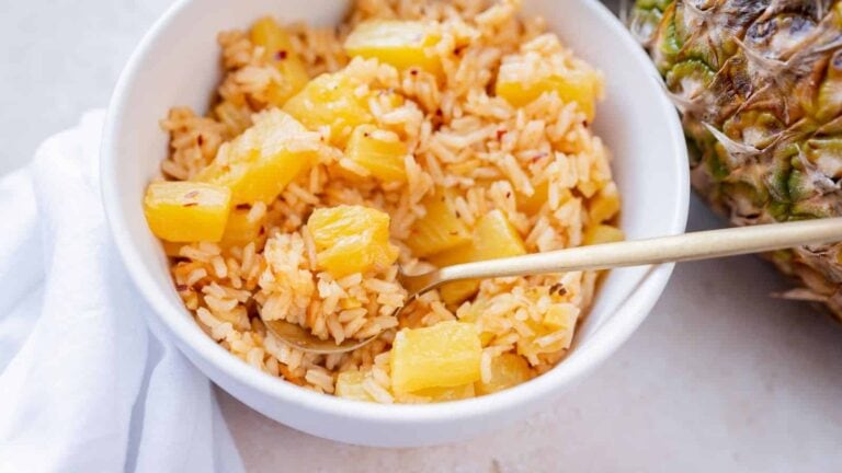 A white bowl filled with rice and pineapple chunks, with a spoon resting inside; a pineapple and a white napkin are beside the bowl.