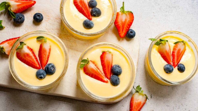 Four glass jars of creamy dessert topped with halved strawberries and whole blueberries, arranged on a white board with extra fruit scattered around.