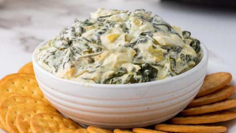 A white bowl filled with creamy spinach artichoke dip, surrounded by round crackers on a white surface.