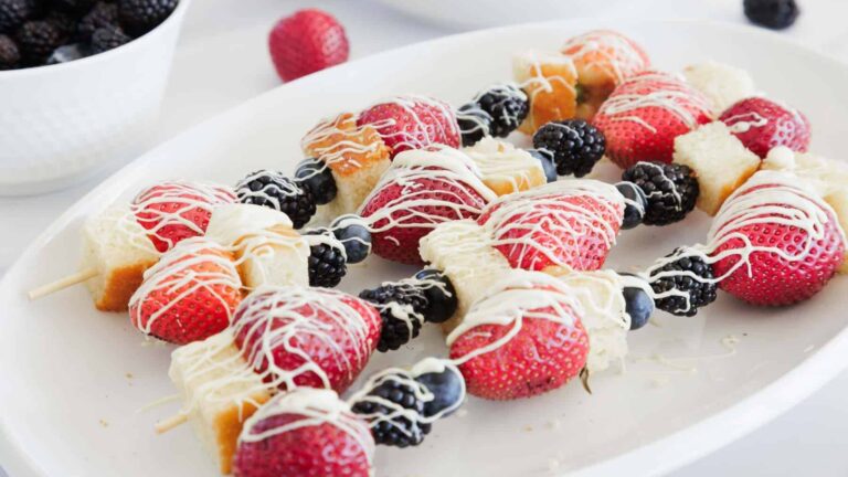 Fruit and cake skewers with strawberries, blueberries, blackberries, and cubes of cake are arranged on a white plate. The skewers are drizzled with white chocolate. A bowl of blackberries is in the background.