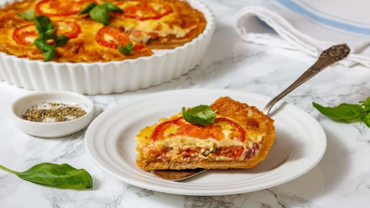 A slice of tomato pie on a white plate with a serving utensil, and a whole tomato pie in a white fluted dish, are shown on a marble surface with a small bowl of spices and basil leaves.