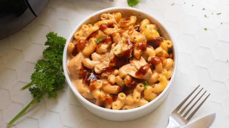 A bowl of macaroni and cheese with pieces of chicken, topped with a drizzle of brown sauce, sits on a white surface beside a fork and sprigs of parsley.
