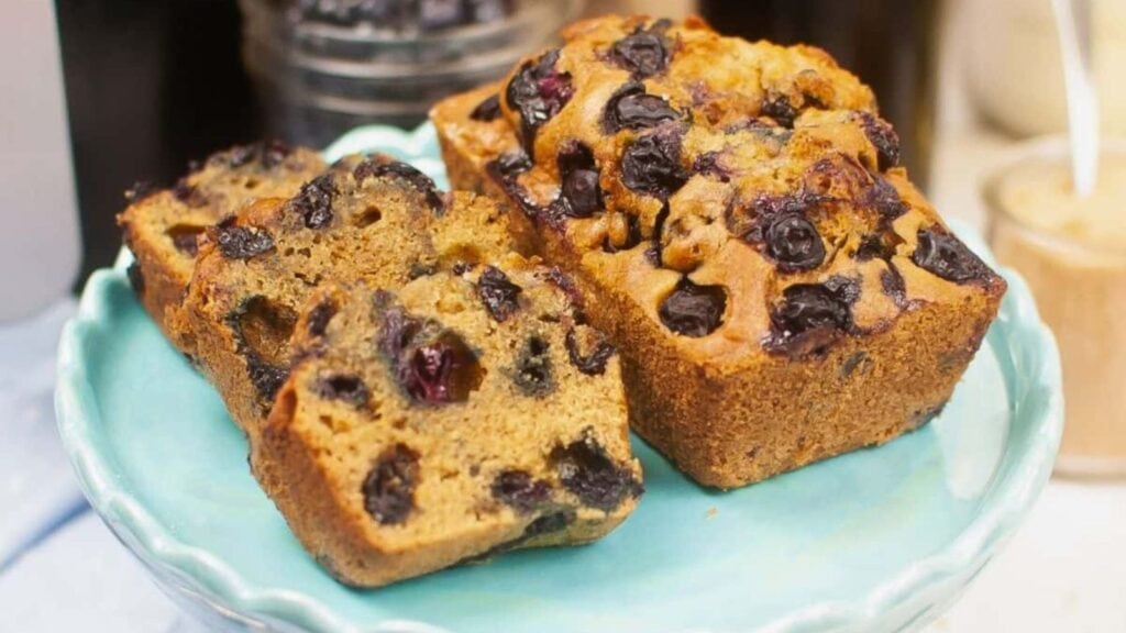 Two slices of blueberry bread on a light blue plate, with a whole loaf behind them and a jar of blueberries in the background.