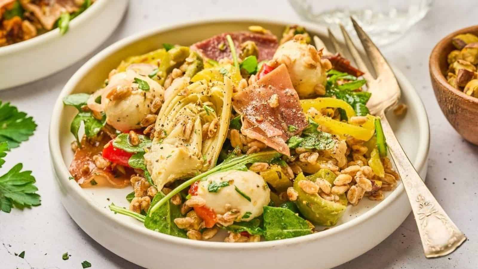 A salad on a plate with mixed greens, mozzarella balls, prosciutto, bell peppers, cherry tomatoes, and grains, garnished with herbs.