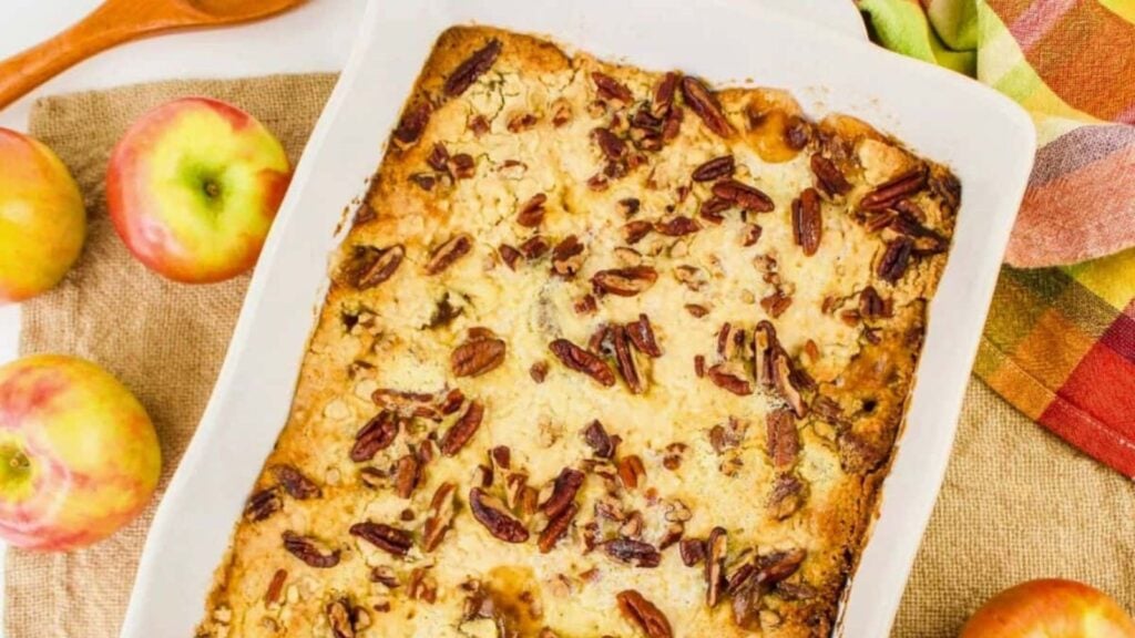 A baked apple dessert topped with chopped pecans in a white dish, surrounded by fresh apples and a colorful cloth.