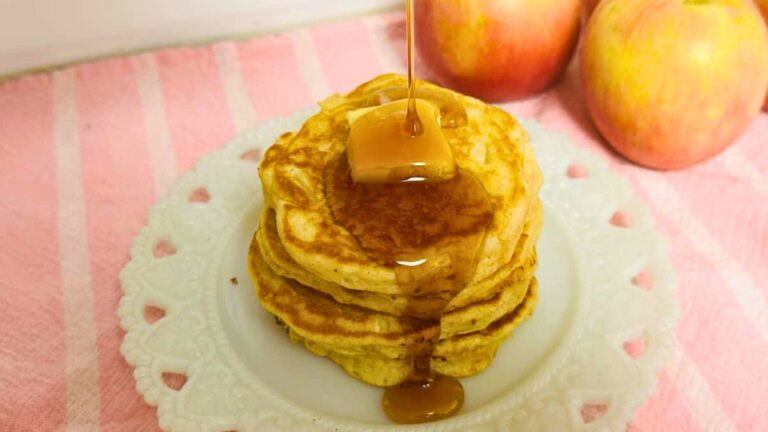 A stack of pancakes topped with a pat of butter is drizzled with syrup, with two apples in the background on a pink striped cloth.