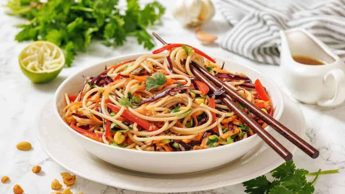 A bowl of Asian-inspired noodle salad with vegetables, garnished with cilantro, chopsticks on top, and ingredients like lime, peanuts, and fresh herbs nearby.