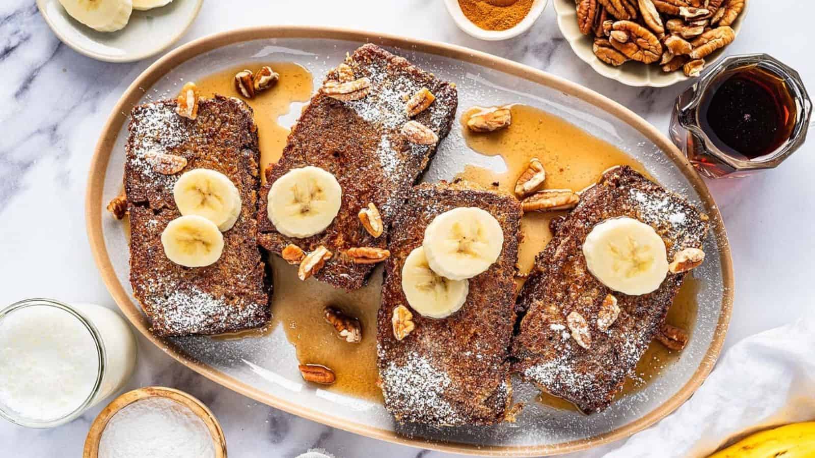 A platter of banana bread french toast with powdered sugar, banana slices, and pecans, drizzled with syrup.