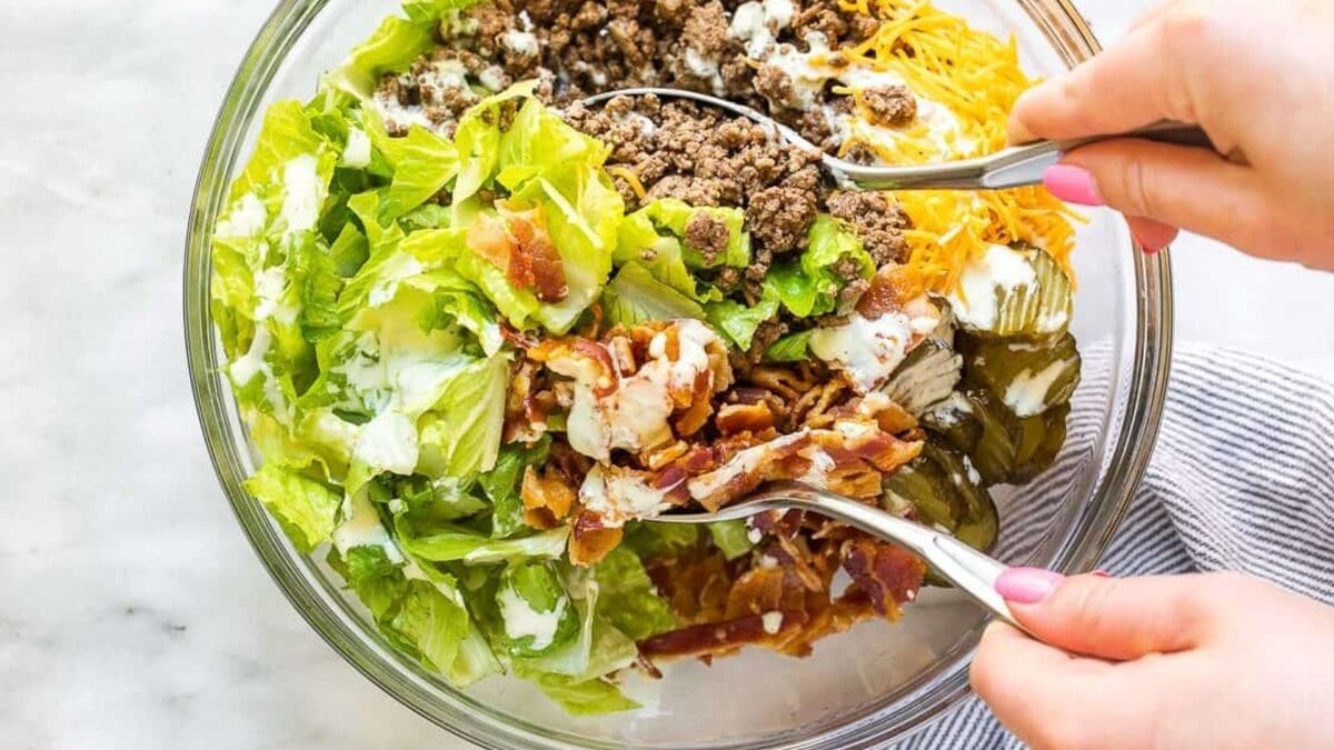 A person holding serving spoons in a bowl of Big Mac salad.