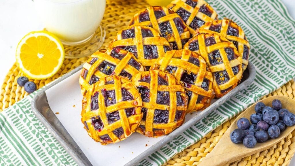 A tray of mini blueberry lattice pies is displayed next to fresh blueberries, a halved orange, and a glass of milk on a woven placemat with a green-striped napkin.