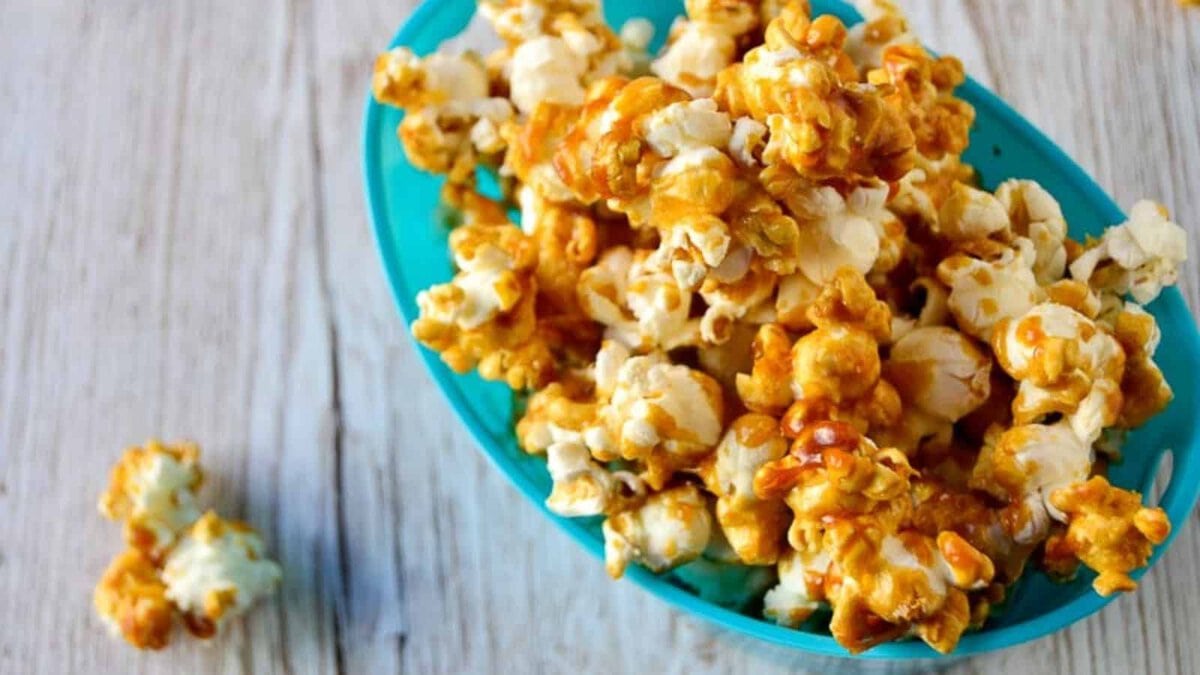 A blue bowl filled with caramel-coated popcorn sits on a wooden surface, with a few pieces of popcorn beside it. This delicious treat is one of the best movie snacks, perfect for your next movie night.