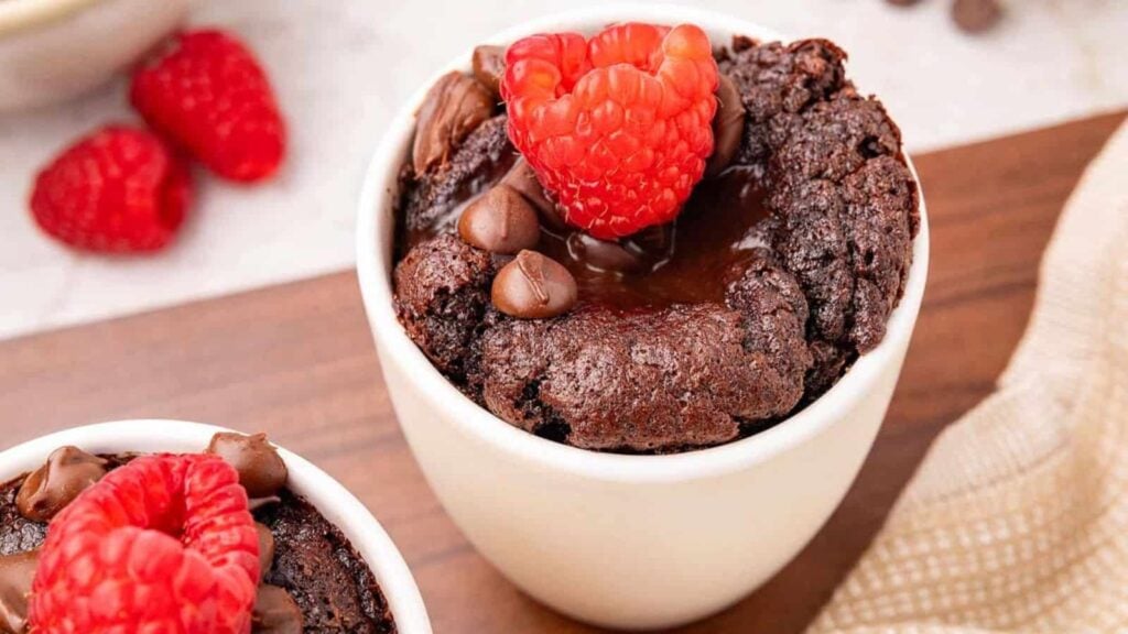 Chocolate mug cake topped with fresh raspberries and chocolate chips in a white cup, placed on a wooden surface.