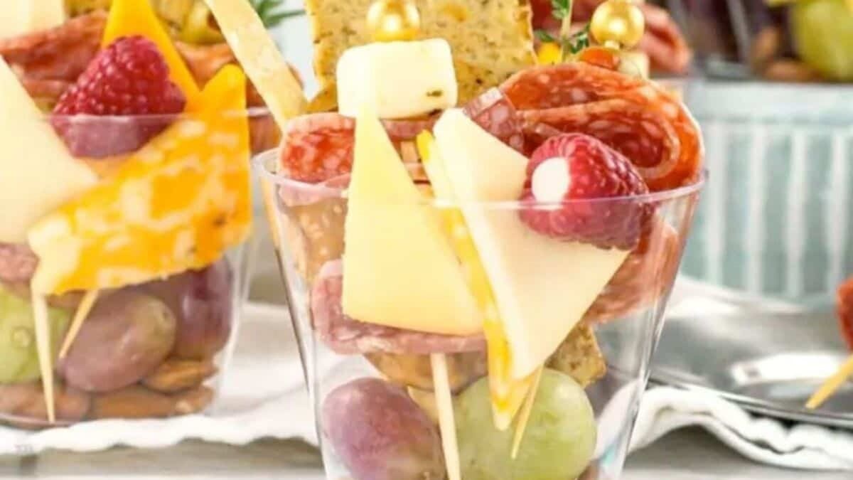 A clear plastic cup filled with grapes, almonds, cheese cubes, salami slices, a raspberry, and crackers, arranged on skewers for a single-serve charcuterie snack.