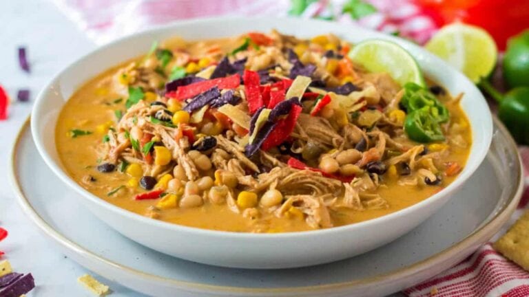 A bowl of creamy chicken chili topped with shredded cheese, tortilla strips, corn, black beans, and lime slices.