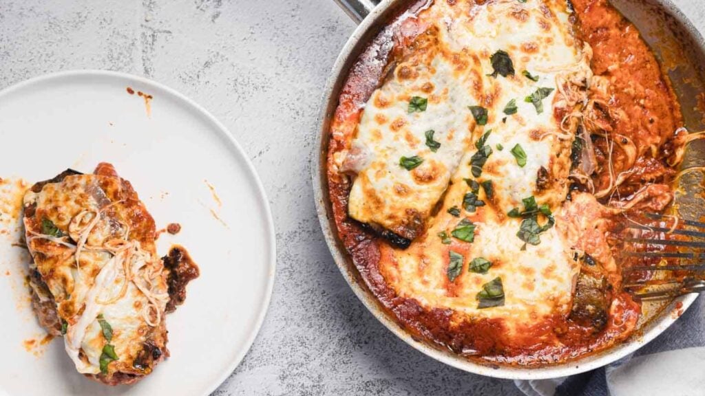 A skillet contains baked eggplant topped with melted cheese and tomato sauce, garnished with fresh basil. A single serving of the dish is plated on a white plate beside the skillet.
