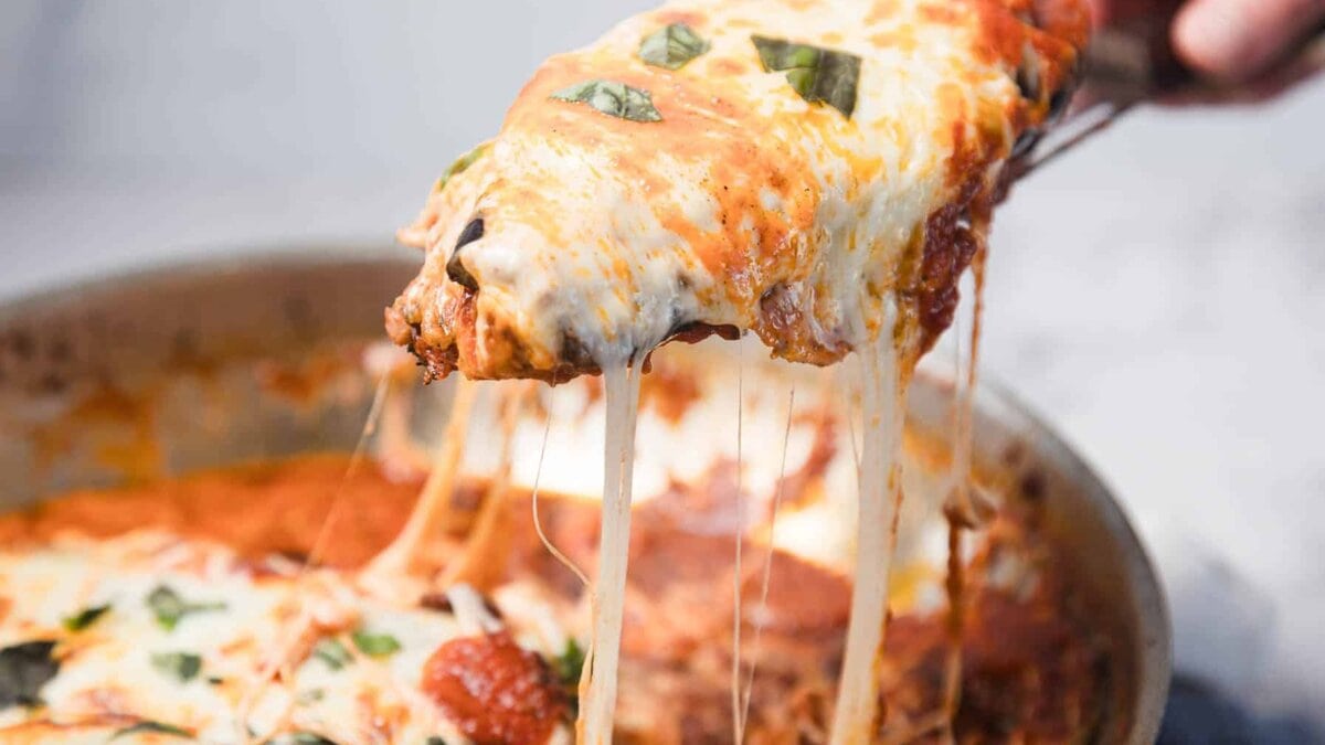 A close-up of a pan of cheesy baked lasagna with a serving being lifted out, showing melted cheese stretching from the pan and bits of fresh basil on top.