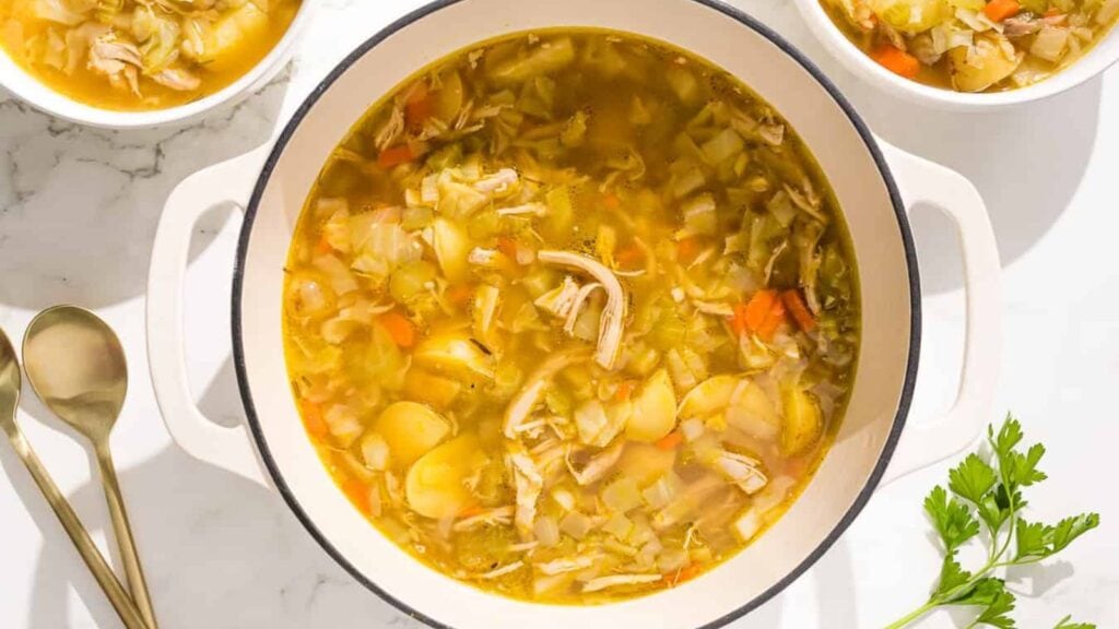 A bowl of chicken and cabbage soup is shown on a rustic wooden cutting board with parsley and a red linen.