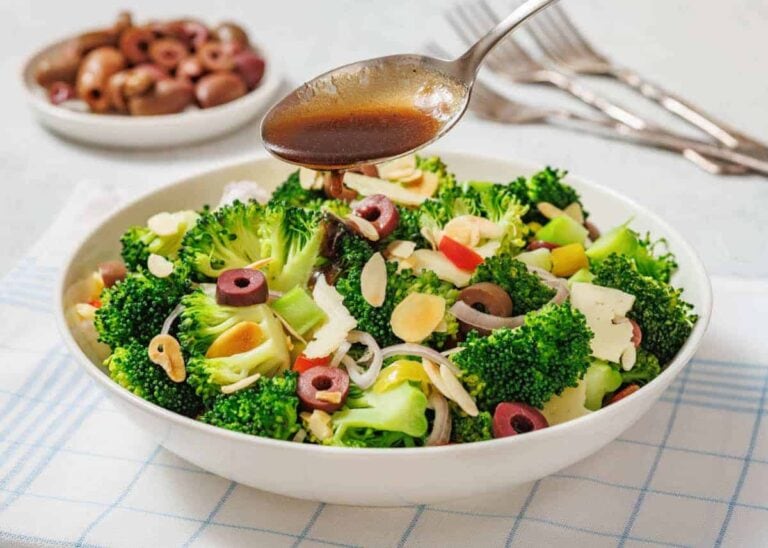 A bowl of Italian broccoli salad with sliced olives, almonds, and vegetables is being drizzled with dressing from a spoon.