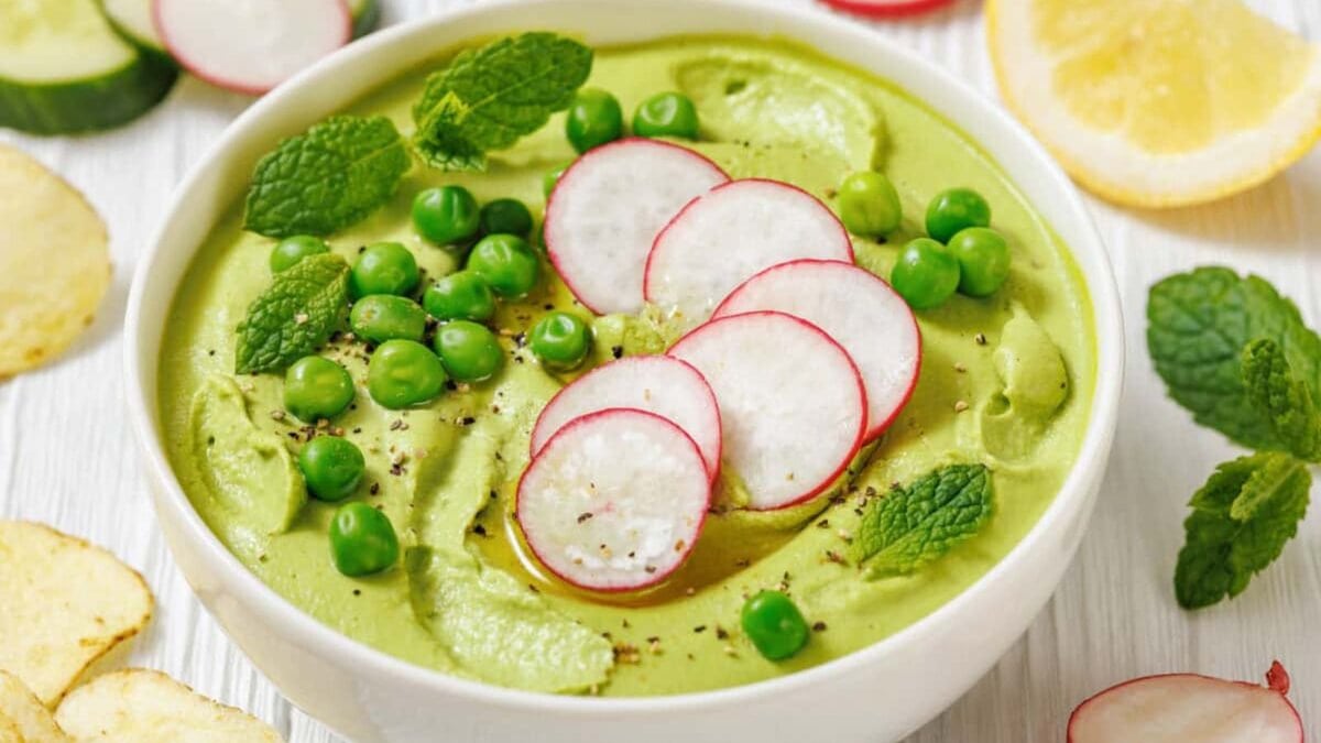 A bowl of Green Pea Hummus with Mint, topped with sliced radishes, green peas, mint leaves, and black pepper, with lemon, cucumber, chips, and radishes on the side.