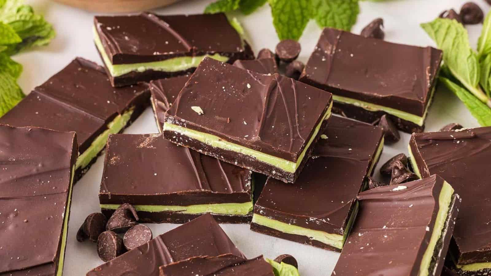 Rectangular mint chocolate bars arranged on a surface, with visible green mint layers between the chocolate. Mint leaves and chocolate chips are scattered around.