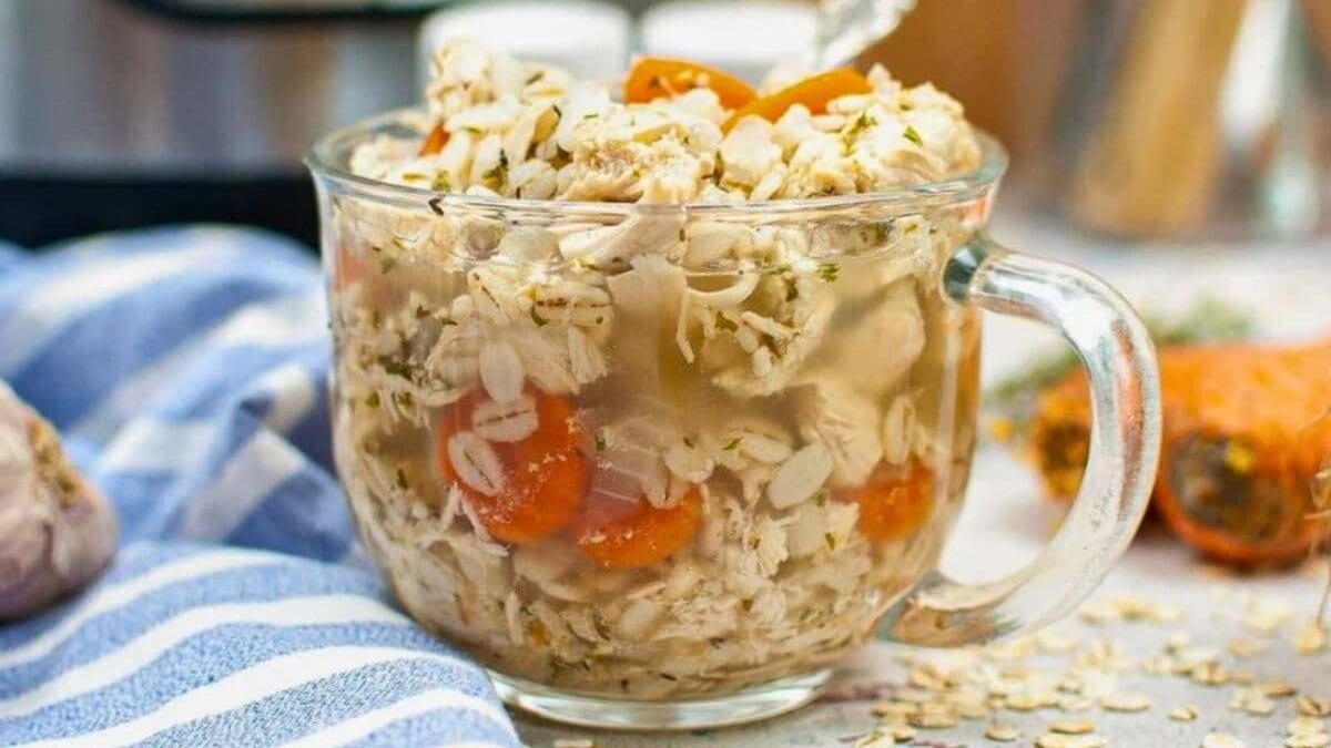 A clear glass mug filled with chicken soup, oats, and carrot slices sits on a countertop with a silver spoon inside.