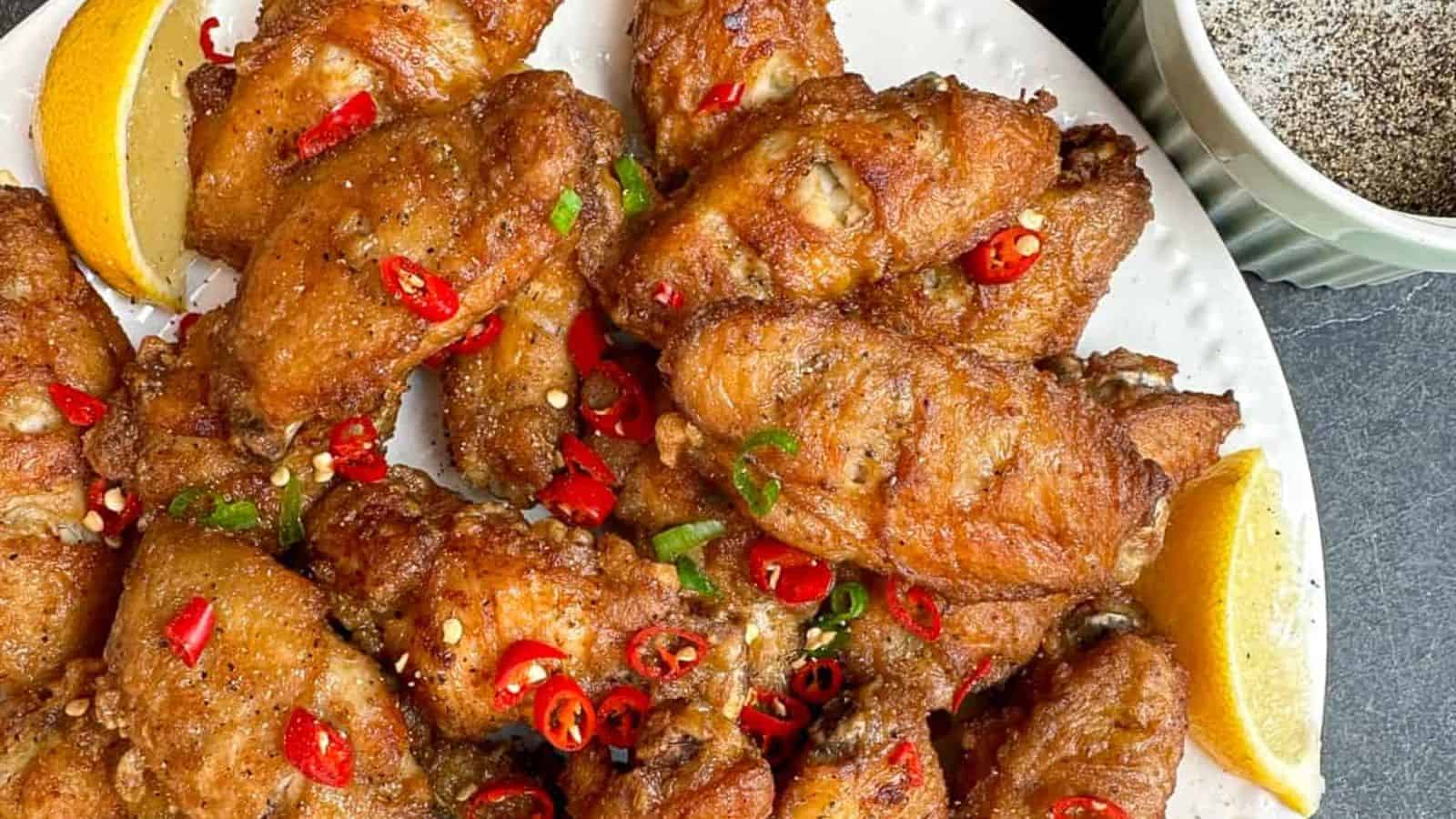 A plate of crispy fried chicken wings garnished with sliced red chilies and green onions, served with lemon wedges, next to a bowl of pepper.