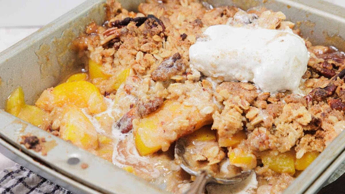 A bowl of peach cobbler topped with melted ice cream sits on a table; a spoonful of cobbler with peaches and crumbly topping is held above the bowl. A checkered cloth is in the background.