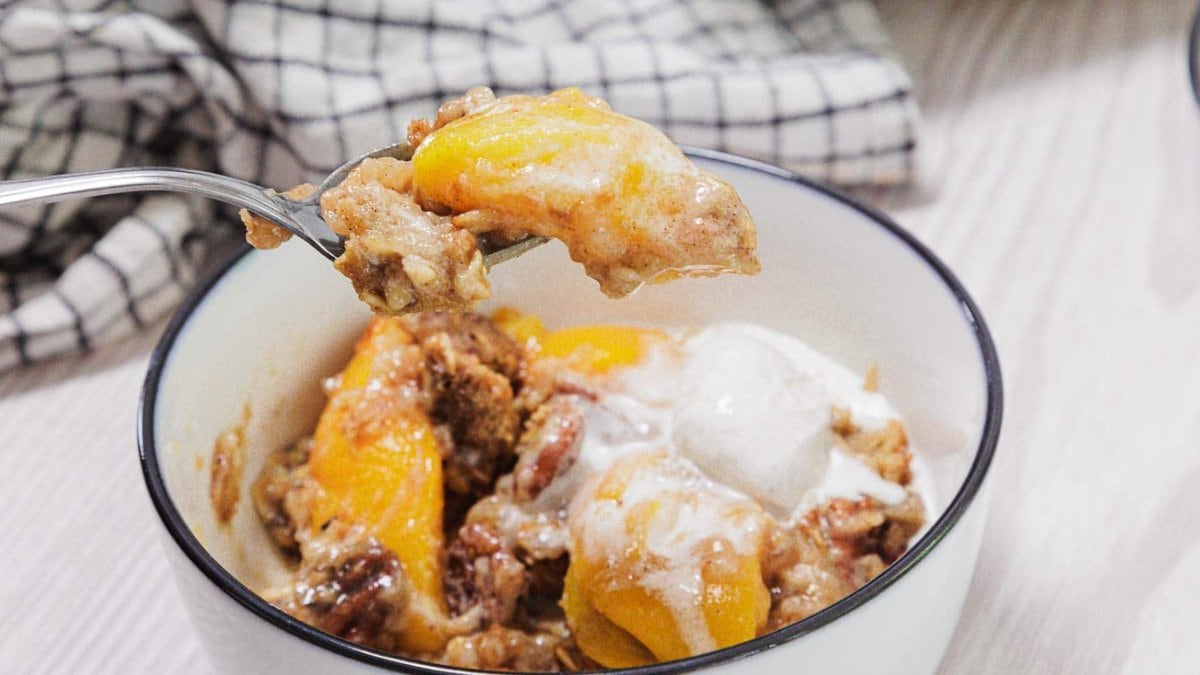 A bowl of peach cobbler topped with melted ice cream sits on a table; a spoonful of cobbler with peaches and crumbly topping is held above the bowl. A checkered cloth is in the background.