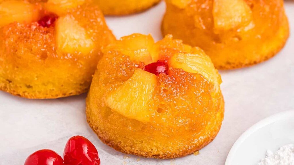 Mini pineapple upside-down cakes topped with pineapple chunks and cherries on a white surface.