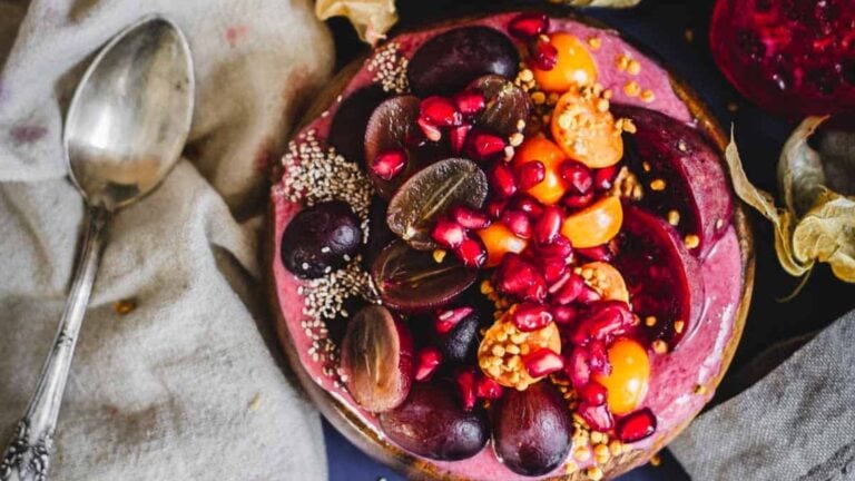 A smoothie bowl topped with halved grapes, sliced plums, pomegranate seeds, golden berries, chia seeds, and bee pollen, placed next to a vintage spoon on a beige cloth.