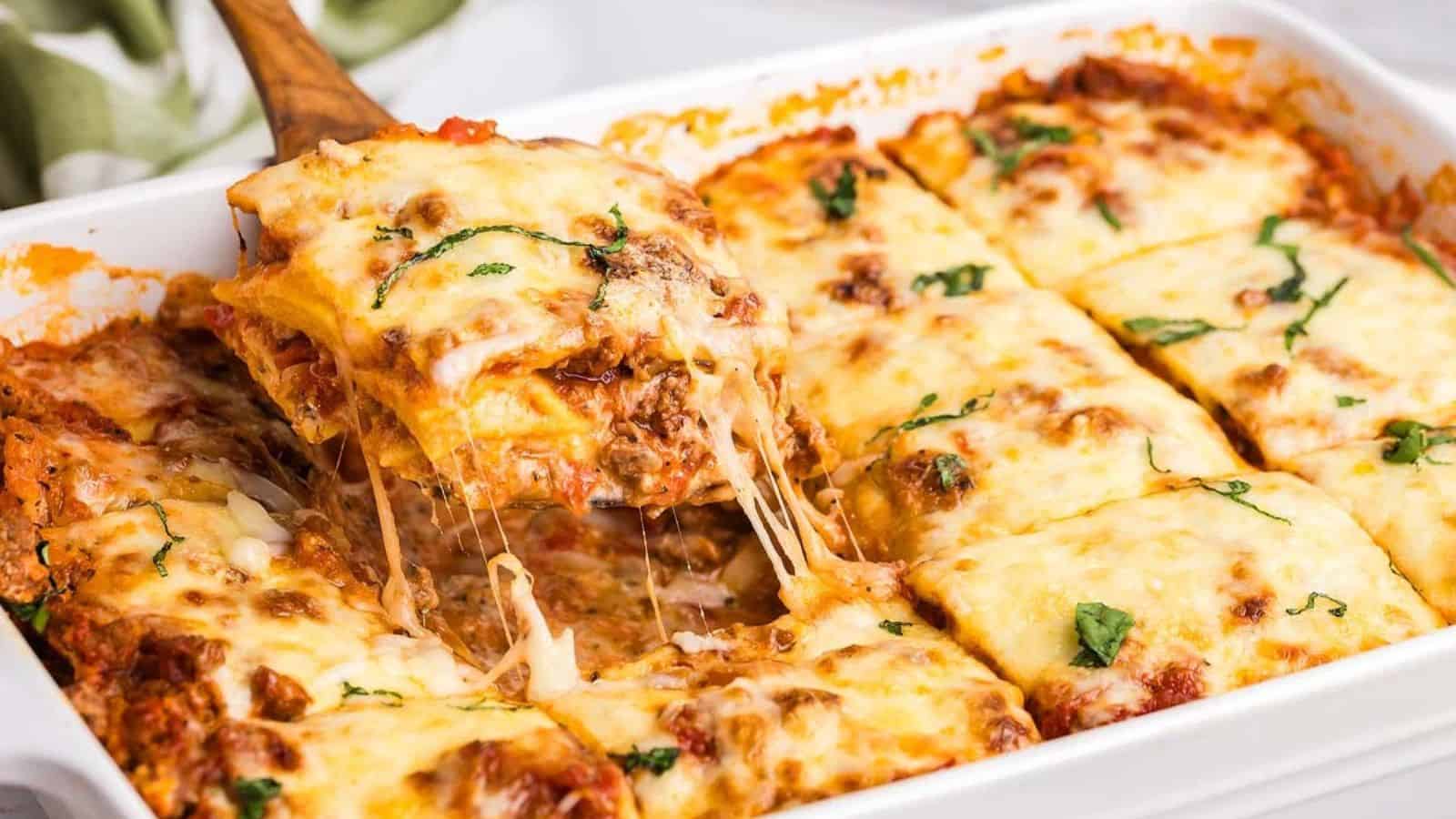 A baked ravioli lasagna in a white dish with cheese and basil on top. A piece is being lifted, showing melted cheese stretching from the dish.