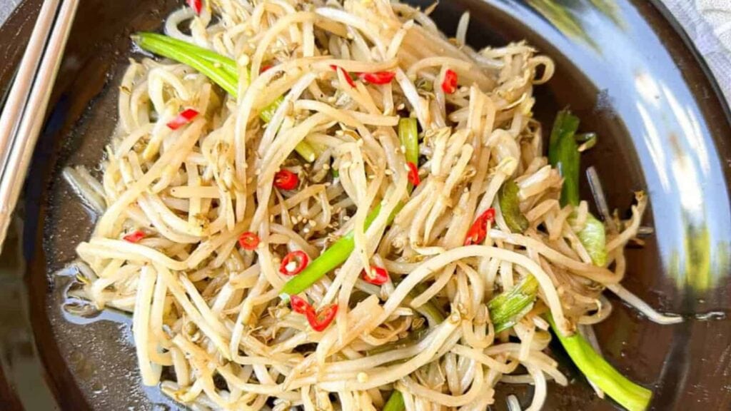 A plate of stir-fried bean sprouts with sliced red chilies and green onions, served on a dark dish with chopsticks.