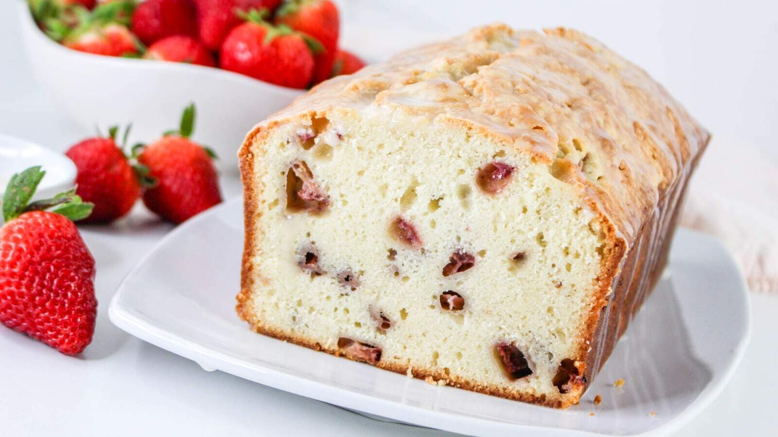 A loaf of strawberry bread on a white plate, with a bowl of fresh strawberries in the background.