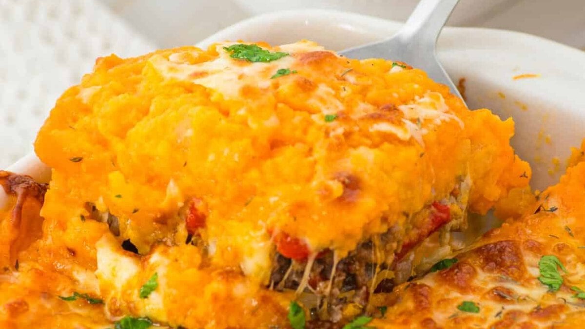 A baked sweet potato casserole topped with melted cheese and chopped parsley, served in a white rectangular dish on a wooden board. Small bowls of fresh herbs and seasonings are visible in the background.