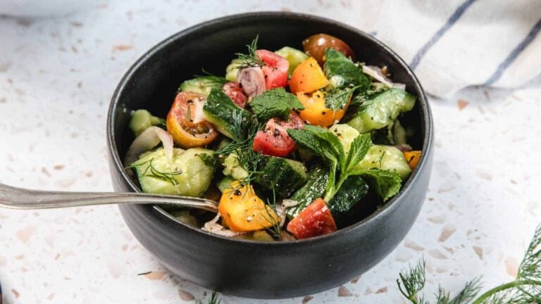 A black bowl filled with chopped tomatoes, cucumbers, herbs, and onions, with a spoon resting inside on a light-colored surface.