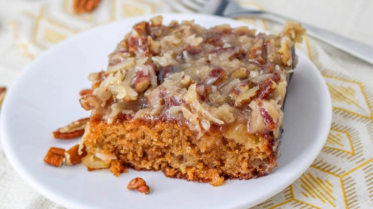 A slice of caramel pecan cake on a white plate, topped with chopped pecans and shredded coconut. A fork is in the background.