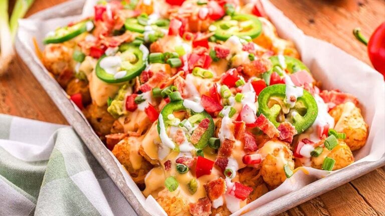A tray of loaded tater tots topped with melted cheese, diced tomatoes, jalapeño slices, bacon bits, green onions, and a drizzle of creamy sauce.