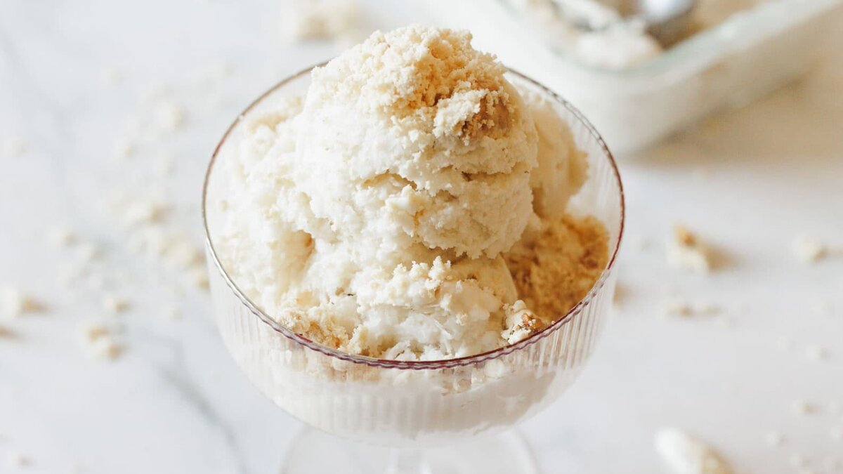 A glass dessert dish filled with scoops of vanilla ice cream, topped with crumbled cookies, sits on a white surface with crumbs scattered around.