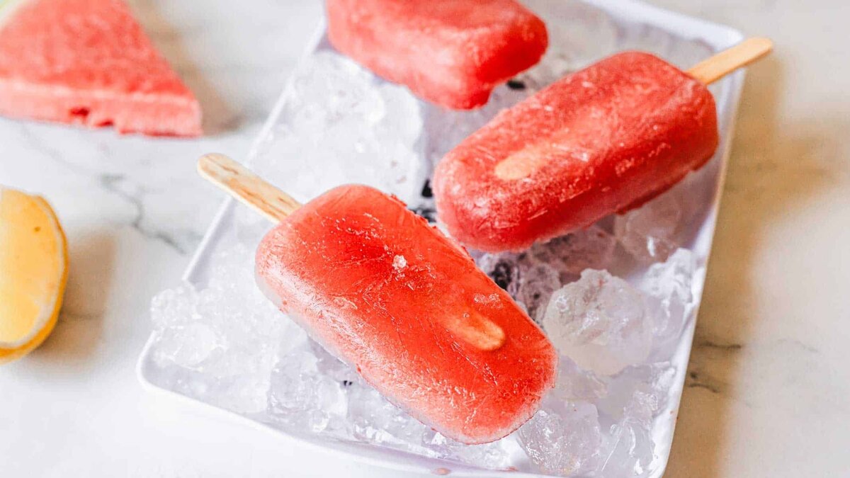 Three red popsicles on wooden sticks rest on a tray filled with ice cubes. A slice of watermelon and a lemon wedge are visible in the background on a white surface.