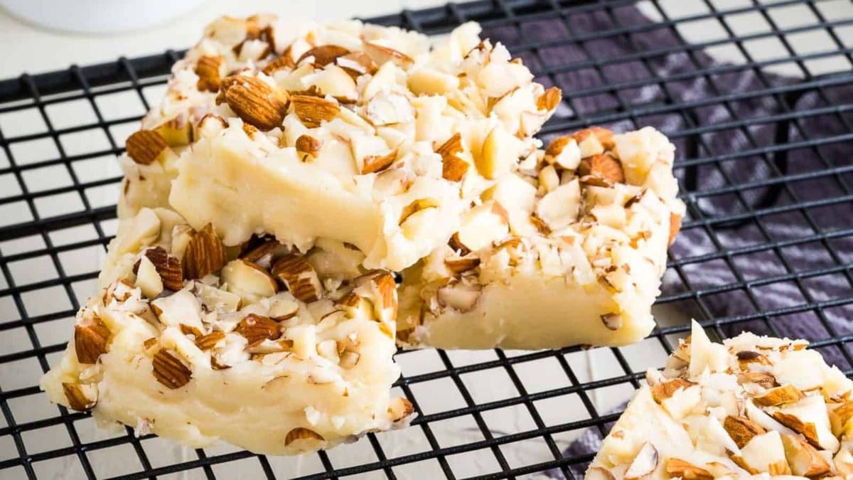 Three pieces of white almond fudge stacked on a black cooling rack, topped with chopped almonds.