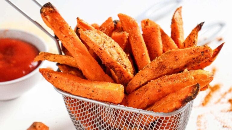 Seasoned sweet potato fries in a metal basket, with a small bowl of red dipping sauce in the background.