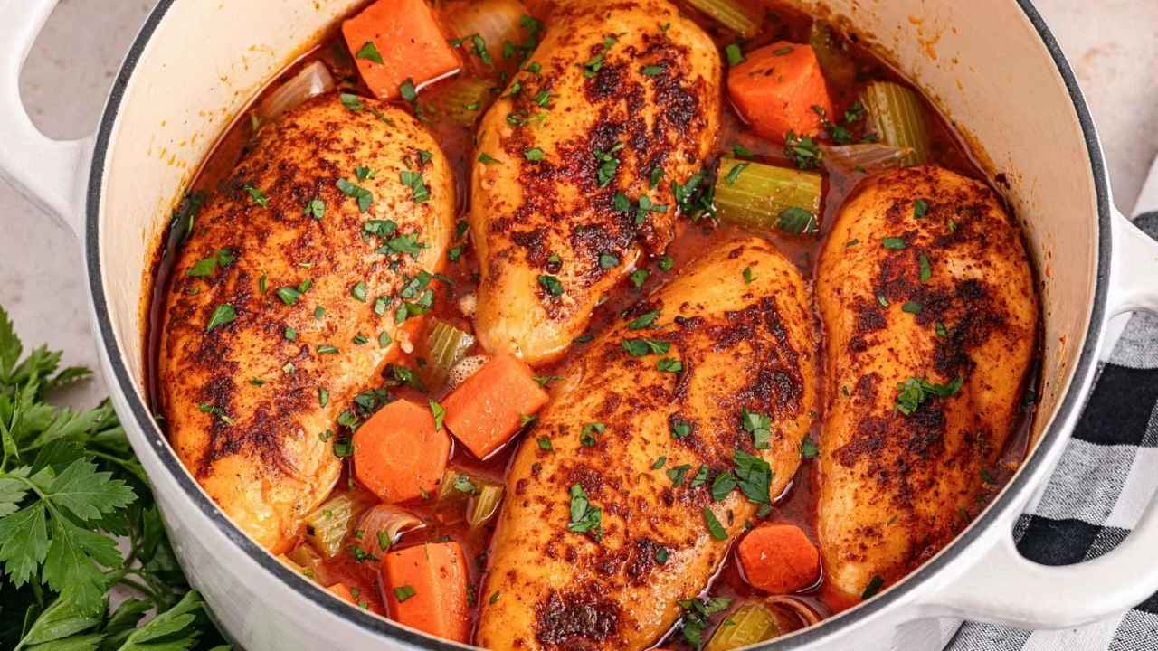 Four seasoned chicken breasts simmer in a pot with chopped carrots, celery, onions, and herbs in a savory broth.