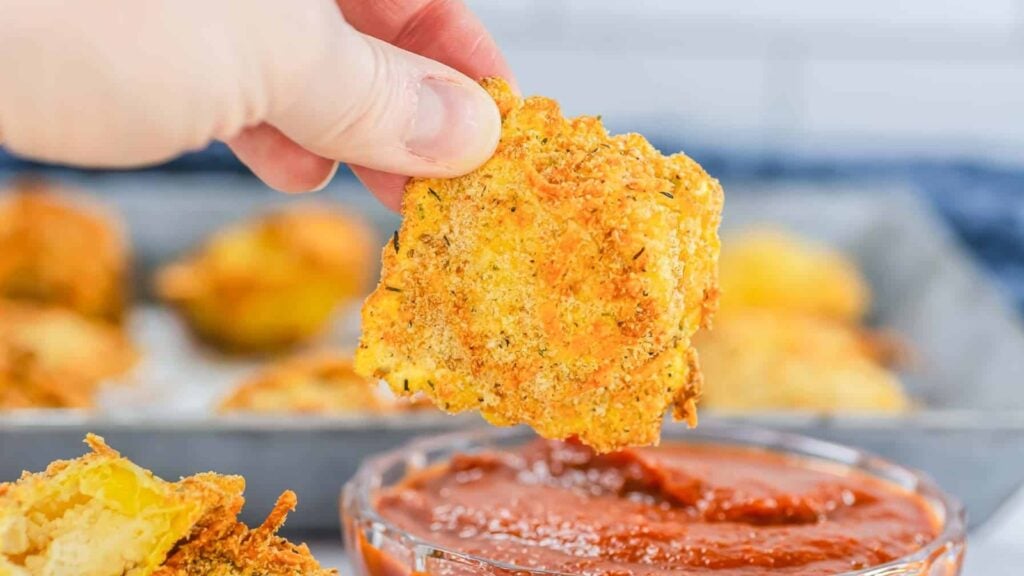 A hand holds a breaded, baked zucchini chip, dipping it into a small bowl of marinara sauce, with more chips visible in the background.