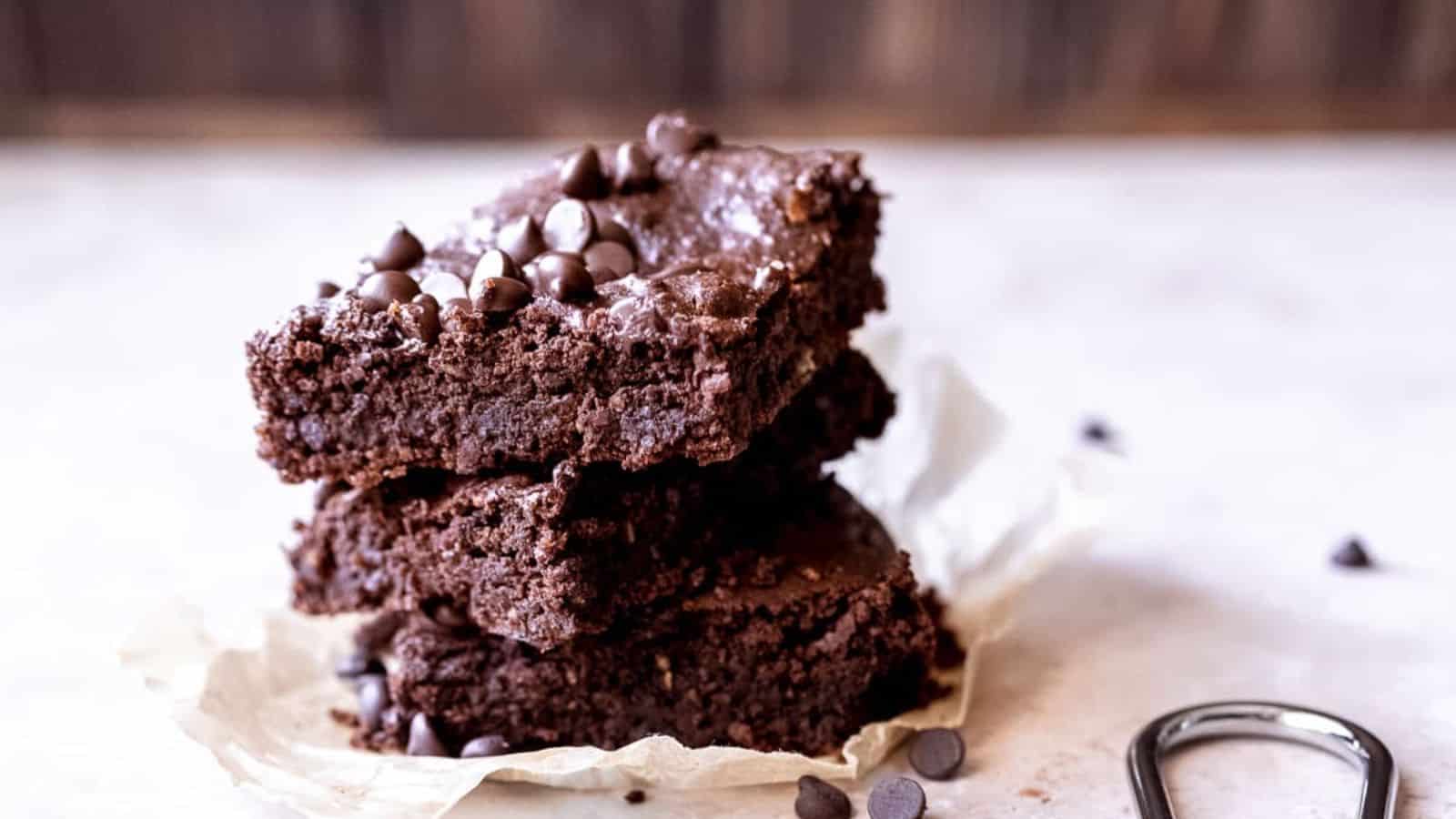 Three chocolate brownies stacked on parchment paper, topped with chocolate chips, with a few chips scattered nearby.