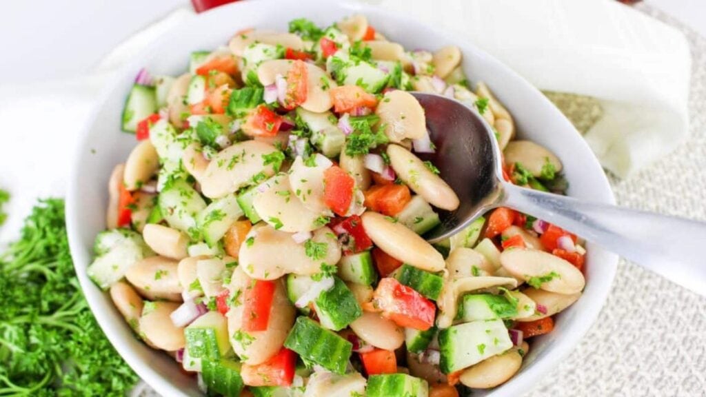 A bowl of bean salad with cucumber, red bell pepper, red onion, and parsley, with a spoon resting inside.