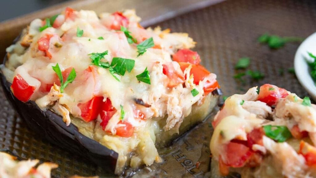 A baked stuffed eggplant half topped with melted cheese, diced tomatoes, shredded meat, and chopped parsley on a baking sheet.