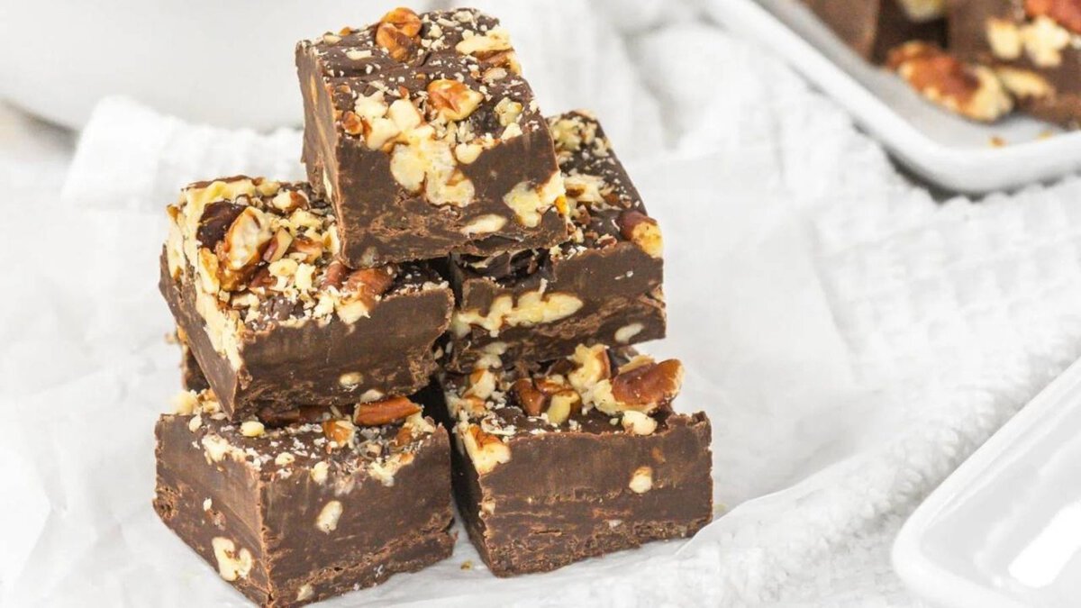 Five pieces of chocolate fudge with chopped nuts are stacked on white parchment paper.
