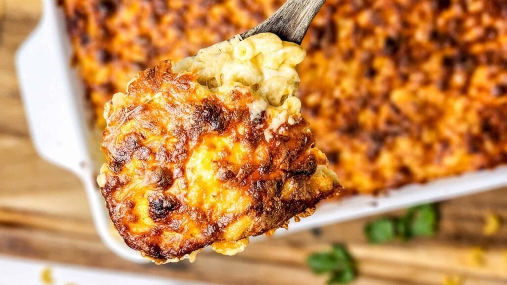 A close-up of a serving of baked macaroni and cheese with a browned, crispy cheese topping, held up by a spoon over a casserole dish.