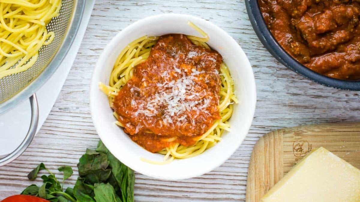 Meaty tomato sauce on a bed of spaghetti noodles in a white background, with a pan of sauce and a bowl of noodles.