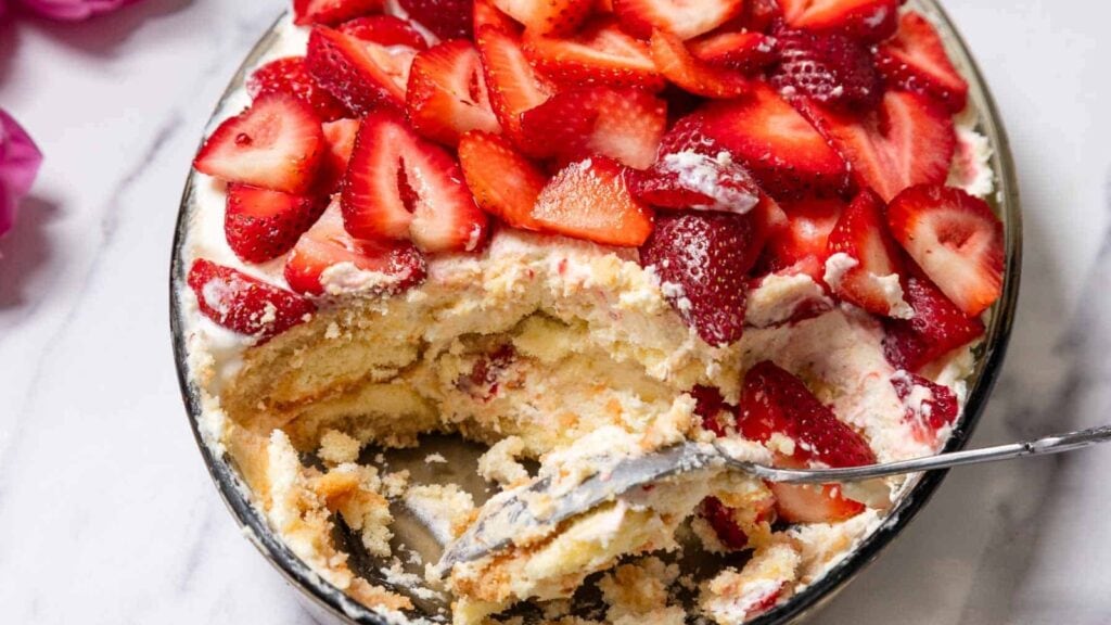 A partially served dish of strawberry tiramisu topped with sliced strawberries, showing layers of cream and cake with a serving spoon inside.